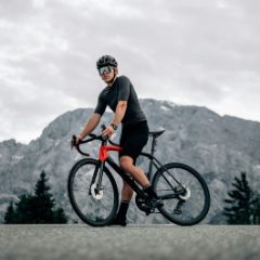 A cyclist in a black helmet standing astride a road bike below mountains