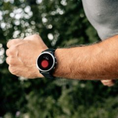 Runner's wrist with a round black sports watch, green trees behind in daylight