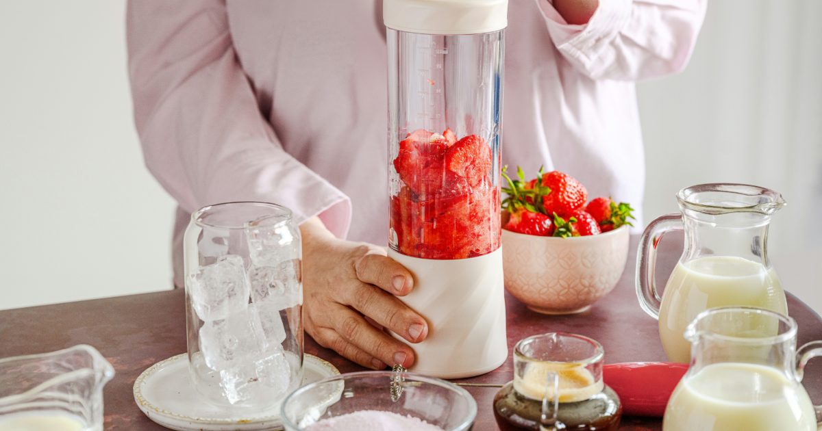 Woman holding a cream personal blender full of strawberries, with a pitcher of ice and bottles of milk on the table