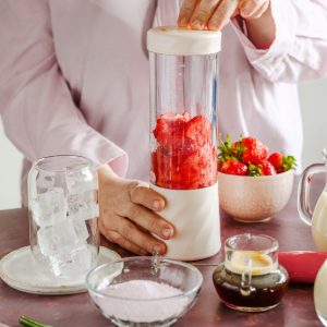 Woman holding a cream personal blender full of strawberries, with a jug of ice and jugs of milk on the table
