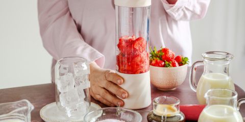 Woman holding a cream personal blender full of strawberries, with a pitcher of ice and bottles of milk on the table
