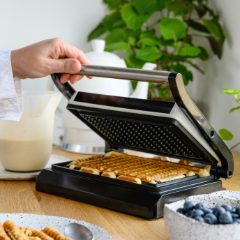Hand lifting the steel lid of a black sandwich maker with waffle plates and golden waffle sticks, in a bright kitchen