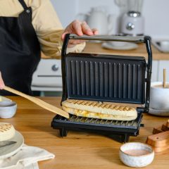 Woman lifting the lid of a small black sandwich grill with a toasted panini inside, on a wooden kitchen worktop