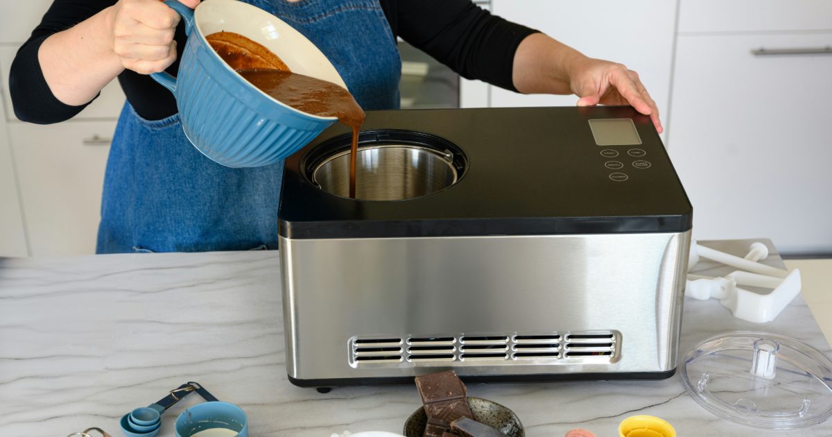 A woman in a denim apron pouring chocolate ice cream mix into a stainless steel ice cream maker on a marble worktop