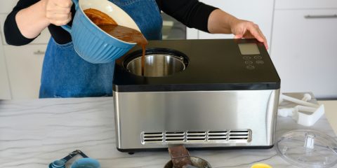 A woman in a denim apron pouring chocolate ice cream mix into a stainless steel ice cream maker on a marble countertop