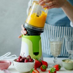 Woman lifting a cup of mango smoothie off a white and lime green personal blender, cherries and strawberries in front