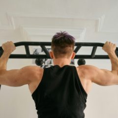 Man in a black vest seen from behind gripping a black multi-grip pull-up bar against a white wall