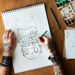 Tattooed hands drawing a cartoon in a spiral-bound sketchbook beside a paint box