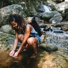 A smiling hiker crouching at a mountain stream with her hands in the water