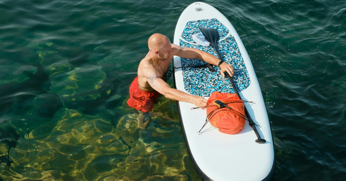 A man standing in clear green water strapping an orange roll-top dry bag to a white paddleboard