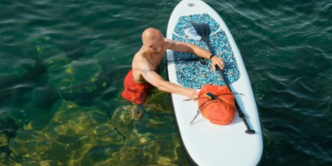 A man standing in clear green water strapping an orange roll-top dry bag to a white paddleboard