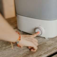 Hand with a beaded bracelet pressing the round dial on the white base of a humidifier with a grey tank, on a wooden table