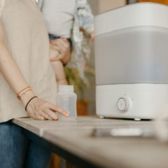 White electric steam steriliser with a frosted body and a round button on its base, on a wooden table beside a woman holding a baby bottle