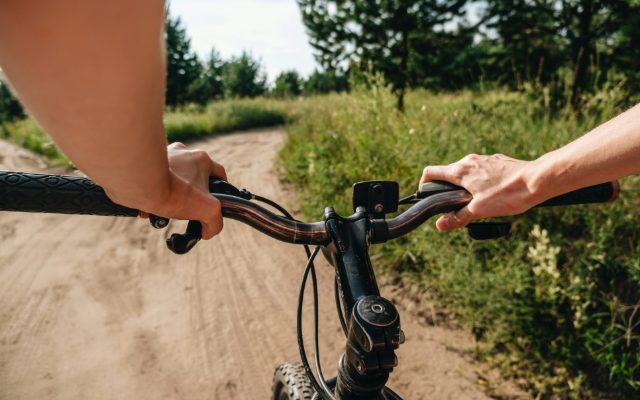 A rider's view of hands on black handlebars riding a sandy path past long grass