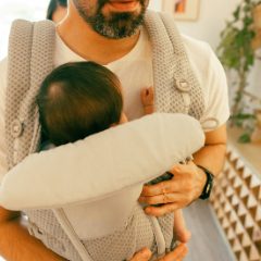 Bearded father in a white T-shirt holding a young baby facing him in a light beige soft front carrier