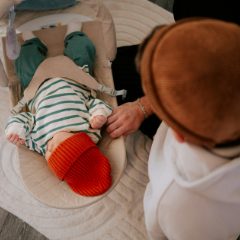 Baby in a striped top and red hat in a beige bouncer with a toy arch, a parent leaning over on a rug