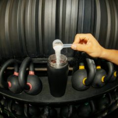 Protein powder being scooped into a black shaker bottle among kettlebells