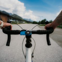 Rider's view over a road bike's drop handlebar with a blue and black twist-lock phone mount in the middle, mountains and a sunny road ahead