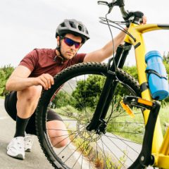 Cyclist in sunglasses and a red jersey crouched checking the front wheel of a yellow bike by a country lane