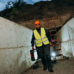 Construction worker in a hi-vis vest carrying a toolbox and a lit portable light through a concrete trench