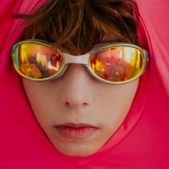 A child wearing orange mirrored swim goggles, framed by a bright pink hood