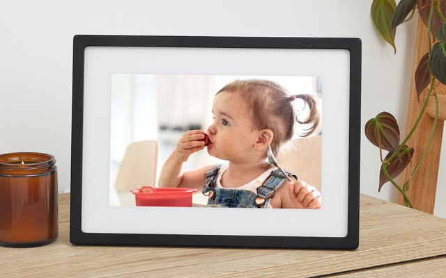 A black digital photo frame with a white mat showing a toddler eating, on a wooden desk beside a candle and a plant