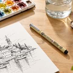 Sakura Pigma Micron fineliners, on an artist's desk beside an ink drawing and a watercolor palette