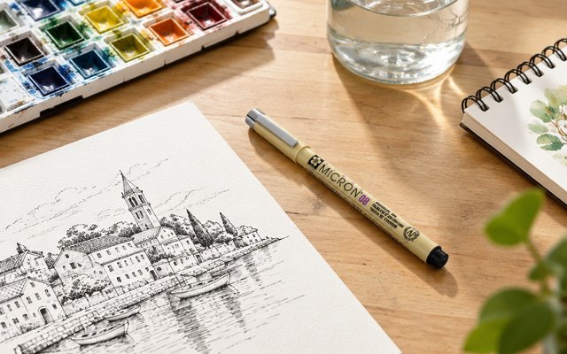 Sakura Pigma Micron fineliners, on an artist's desk beside an ink drawing and a watercolor palette
