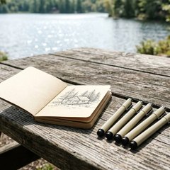 Beige fineliner pens beside a small sketchbook with an ink drawing of a cabin, on a picnic table by a lake