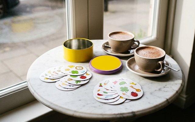 Round symbol cards in two piles on a marble café table beside an open yellow tin, its purple-rimmed lid and two hot chocolates