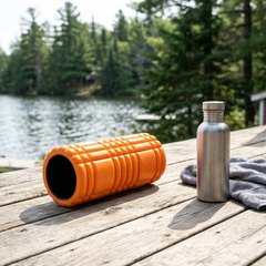 Short orange grid foam roller on a sunlit cottage deck by a lake, beside a steel water bottle and a grey towel