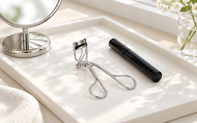 Eyelash curler lying on a white vanity tray beside a mascara tube and a small round mirror, morning light