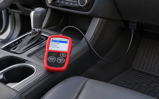 Handheld OBD2 scanner, resting between the front seats of a family SUV, its cable leading to the port under the dashboard, its screen showing a simple menu