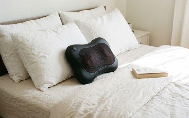 Black and grey massage pillow resting among white pillows on a made bed, a book beside it