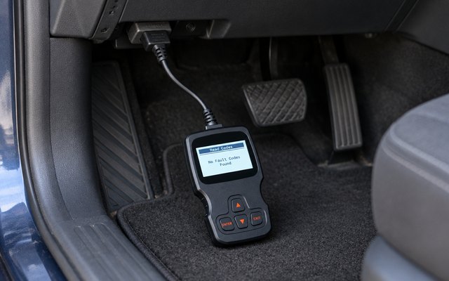 Handheld OBD2 code reader, its cable plugged into the port under a car's dashboard, the reader resting in the driver's footwell, its screen showing a simple reading