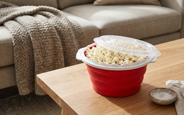 Red collapsible microwave popcorn popper full of popcorn on a wooden coffee table in a bright living room