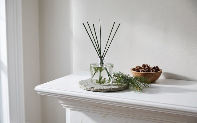 A glass reed diffuser printed with green pine needles, with green reeds, on a stone coaster on a white mantel beside an evergreen sprig and pine cones