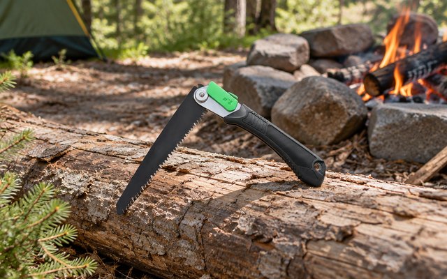 Mossy Oak folding saw, resting on a log by a campfire ring at a wooded campsite