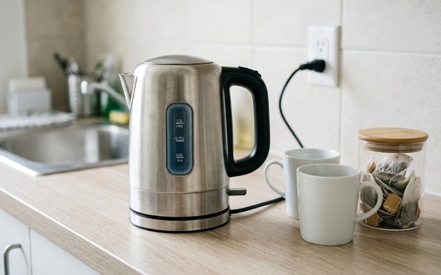 Small brushed-steel electric kettle on an office kitchenette counter beside two white mugs and a jar of tea bags