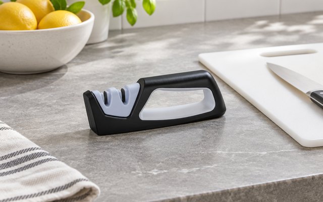 Handheld two-stage knife sharpener, on a gray stone countertop beside a white cutting board and a bowl of lemons