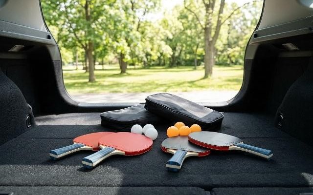 Four table tennis paddles and balls on a car trunk floor beside two zippered bags