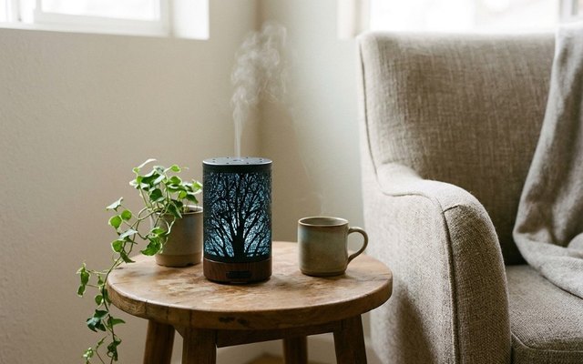 Black metal diffuser with a tree cut-out pattern glowing blue on a side table beside an armchair