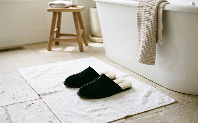Black suede backless slippers with a cream wool lining on a white bath mat beside a freestanding tub