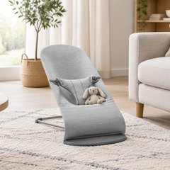 BabyBjörn Bouncer Bliss, baby bouncer on a soft rug in a bright, calm living room beside a sofa, a small knitted toy on its seat
