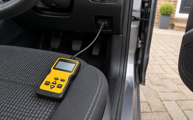 MOTOPOWER OBD2 scanner, resting on the driver's seat of a car parked in a driveway, its cable plugged into the port under the dashboard