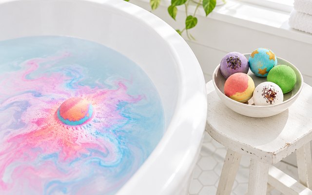 A large bath bomb fizzing in a white tub with more colorful bath bombs in a bowl beside it