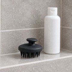 Black silicone shampoo brush on a tiled shower shelf beside a plain bottle