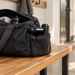 A small massage gun tucked in the side pocket of an open gym bag on a wooden hallway bench
