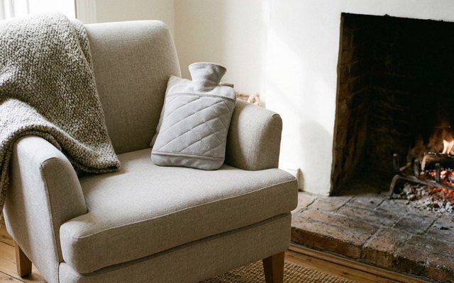 Hot water bottle in a grey quilted cover with a hand pocket, on an armchair by a lit fireplace