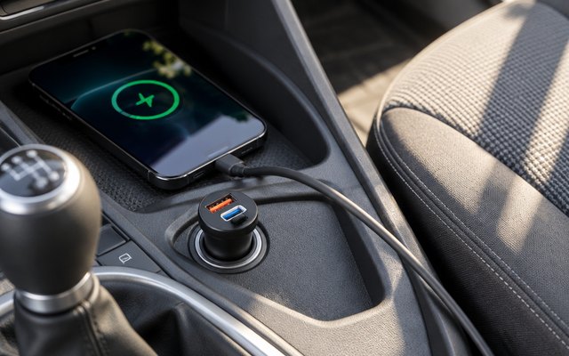 INIU dual-port car charger, sitting flush in the socket of a small city car's centre console, one cable running to a phone on the tray, sunlight across the seat